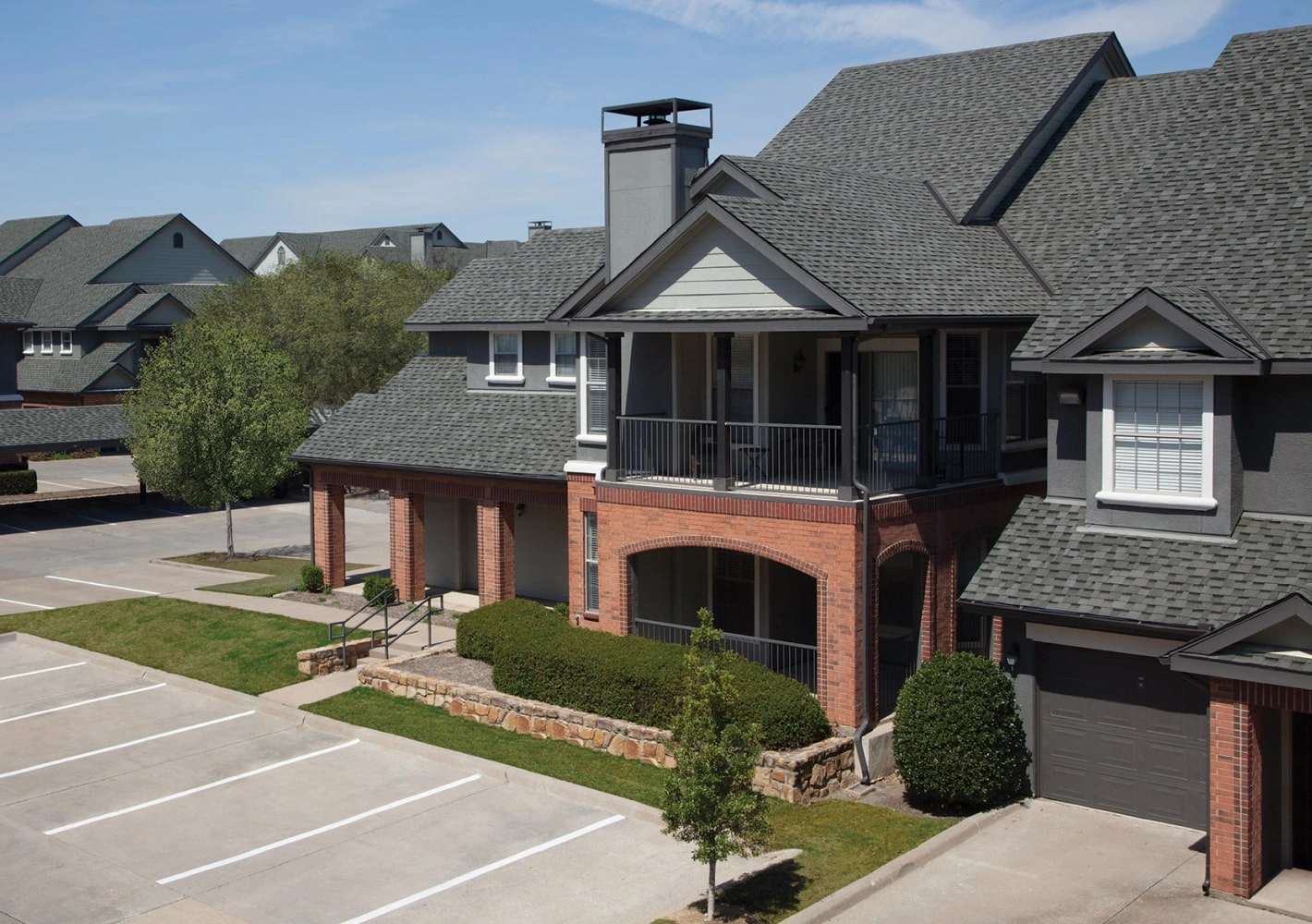A charming two-story residential building in Cambridge, featuring cool colors with red brick and gray siding. It boasts a balcony and chimney, plus it's adjacent to a parking area surrounded by neatly trimmed bushes and trees.