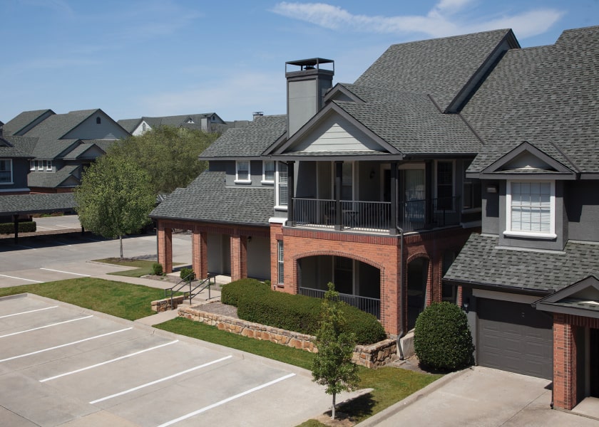 A charming suburban townhouse complex in Cambridge showcases cool colors with its brick and gray siding. It features a covered balcony and an empty parking area, plus a few trees that stand out on this sunny day.
