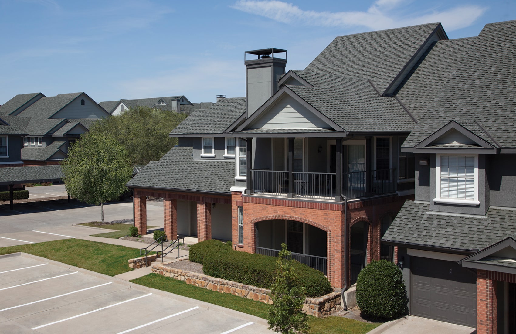 A charming two-story residential building in Cambridge, featuring cool colors with red brick and gray siding. It boasts a balcony and chimney, plus it's adjacent to a parking area surrounded by neatly trimmed bushes and trees.