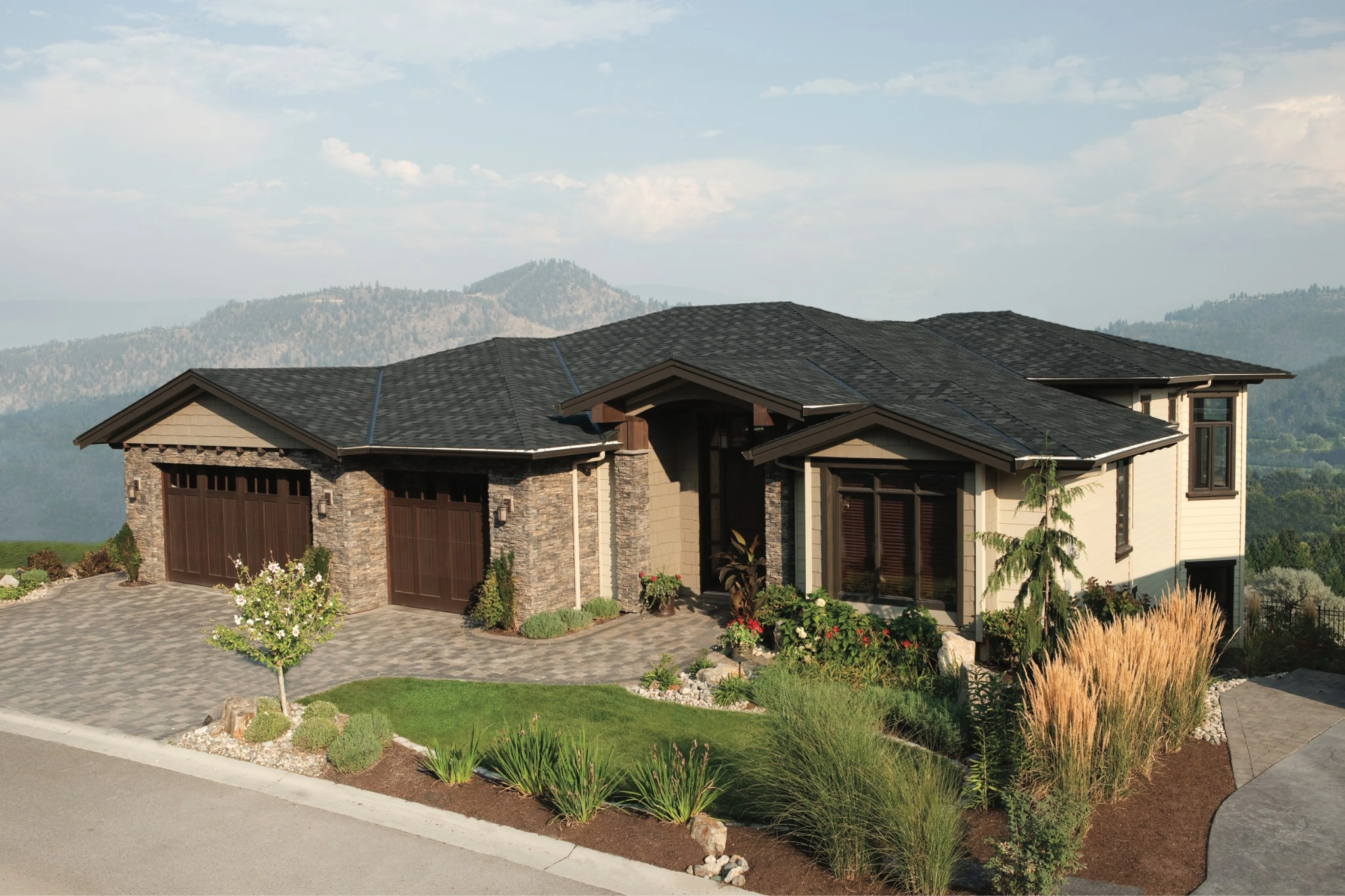 Modern house with stone and wood facade, featuring an Armourshake roof in Shadow Black, three-car garage, landscaped yard, and a breathtaking mountainous backdrop.
