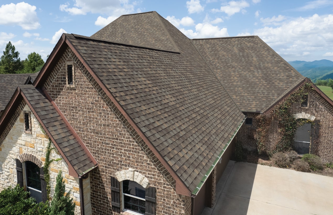 A brick house with a steep, Weatherwood shingled roof. The structure features arched windows, a garage, and lush greenery. A blue sky with clouds graces the background.