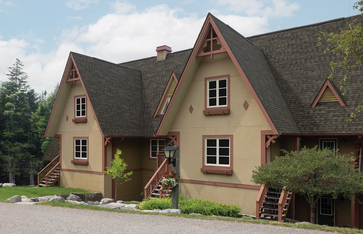 Chalet-style building with three gabled roofs, wooden trim, and beige siding exudes a timeless dynasty appeal. A welcoming pathway winds through lush greenery in front.