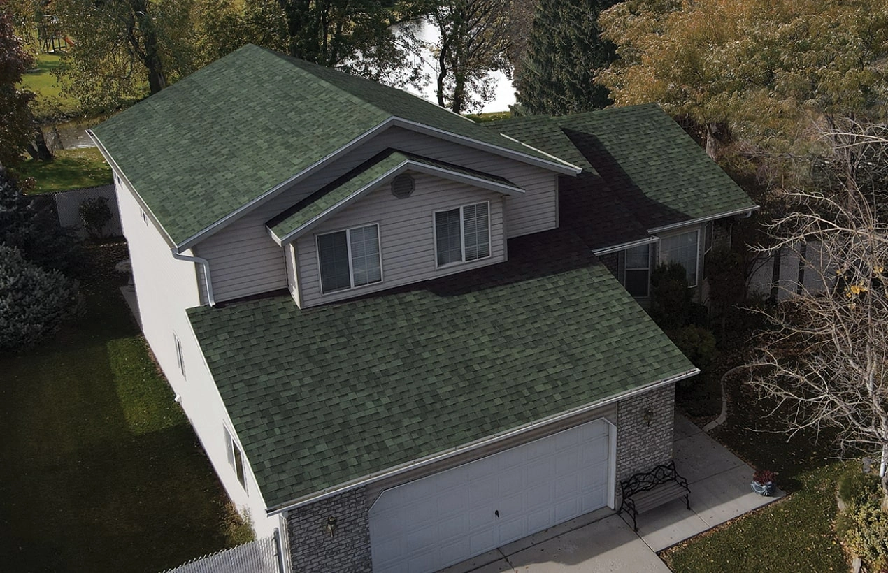 An aerial view reveals a charming two-story house with an emerald green shingle roof, white siding, and brick accents, nestled gracefully amidst a dynasty of towering trees.