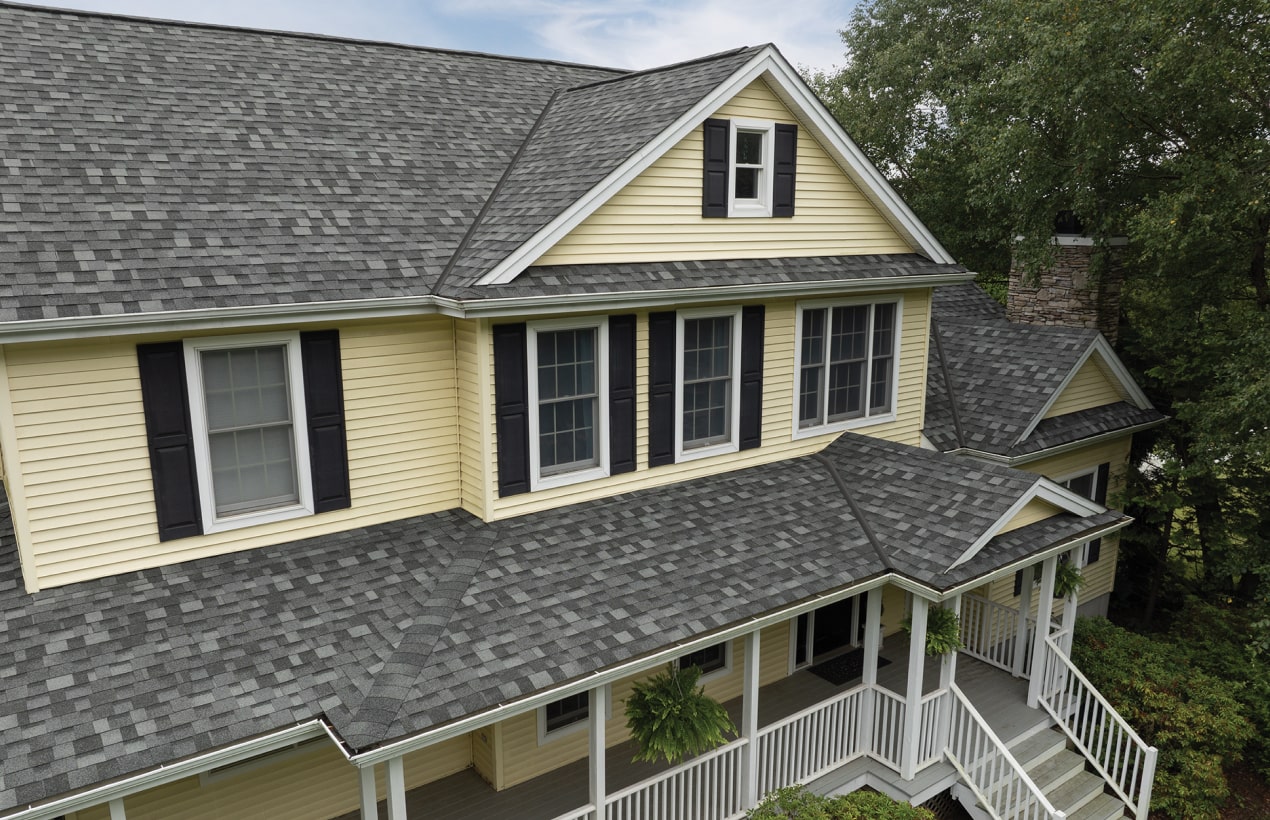 A yellow house with Summit Grey shingles, black shutters, and a Nordic-inspired wraparound porch. There are trees in the background and potted plants hanging on the porch.