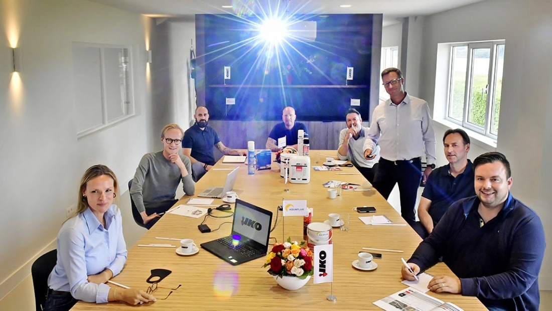 Eight people sitting around a conference table with notepads and laptops. A projector shines brightly.