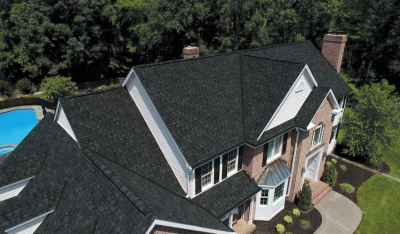 Aerial view of a large brick house with dark shingle roofing, surrounded by trees and featuring a pool in the backyard.
