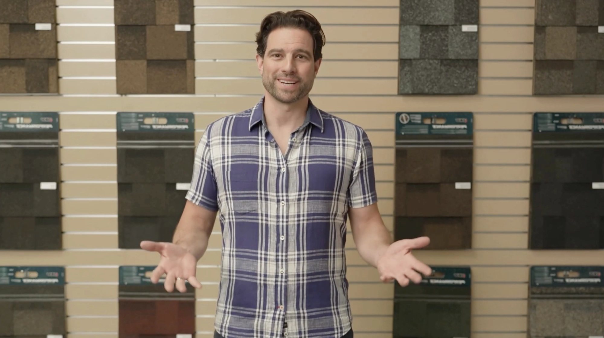 A person stands in front of a wall display of roofing materials, wearing a blue and white plaid shirt, gesturing with open hands like real estate expert Scott McGillivray discussing home renovation tips.