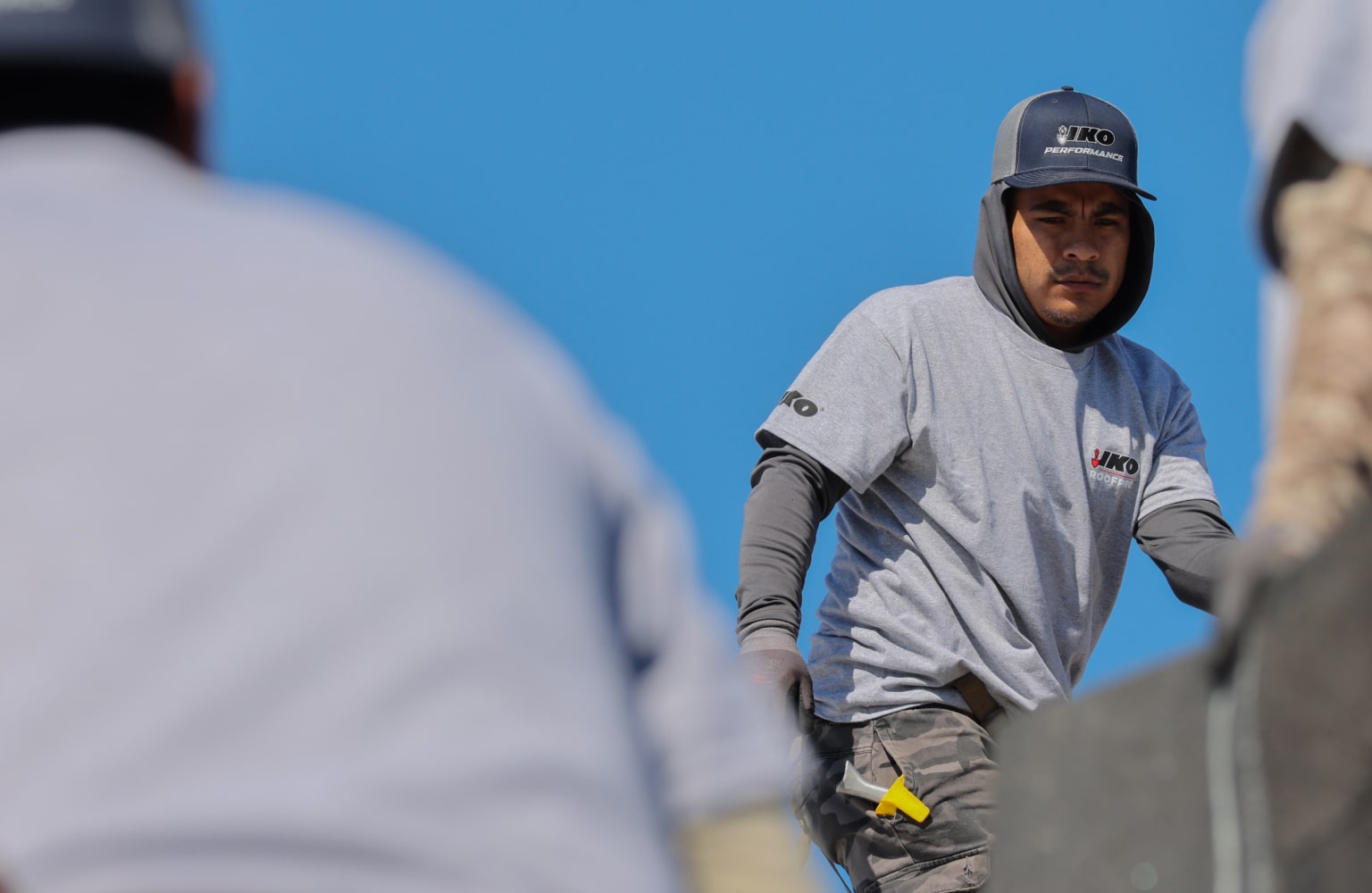 A construction worker in a gray uniform and cap, holding tools, stands outdoors with a clear blue sky in the background.