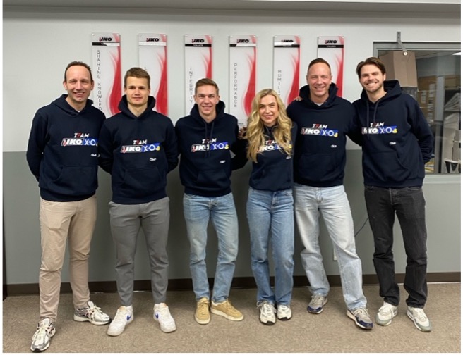 Six people stand indoors, wearing matching black hoodies with text, embodying the spirit of Team IKO X2O. Positioned confidently in front of wall-mounted displays, they reflect the dedication and unity found at events like the ISU World Cup and within organizations such as IKO North America.