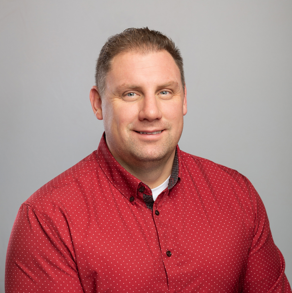 A person wearing a red polka dot shirt, smiling, with short hair, poses against a plain gray background.